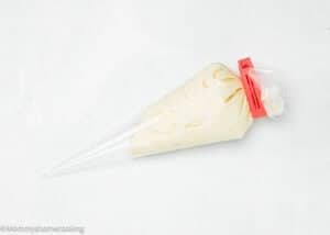 A clear piping bag filled with creamy white frosting, sealed at the top with a red bag clip, is placed on a white surface.