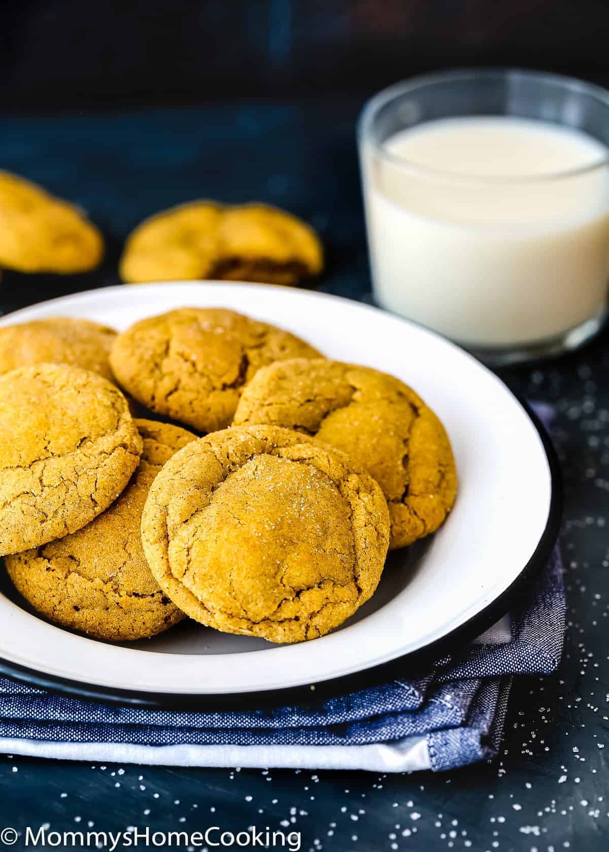 A plate of golden brown cookies sits on a stack of napkins beside a glass of milk, with more cookies in the background.