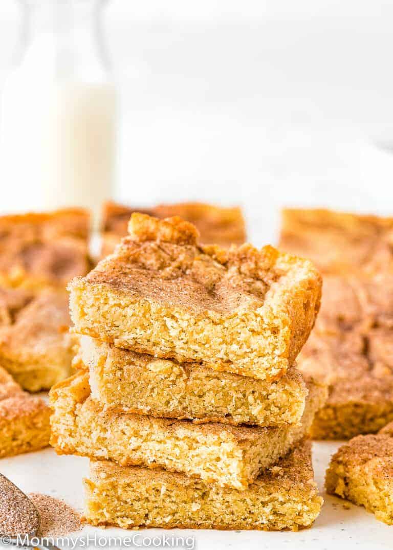 A stack of three golden brown snickerdoodle cookie bars sits in front of more bars, with a bottle of milk blurred in the background—perfect for anyone searching for irresistible Egg-Free Bar Recipes.