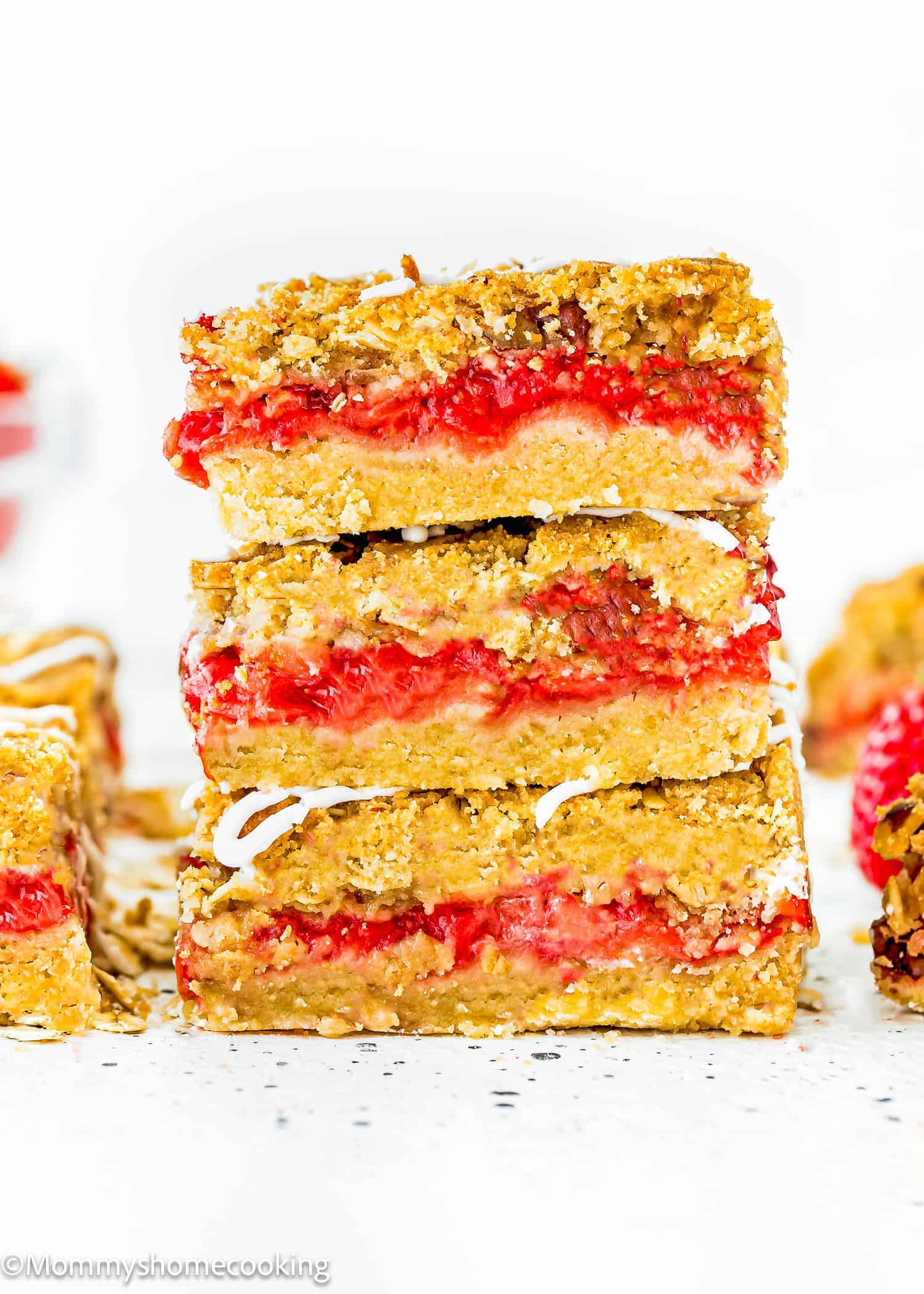 Three stacked oat crumble bars with a red fruit filling and drizzle on top, perfect for anyone searching for 10 Easy Egg-Free Bar Recipes Every Baker Needs, with more bars and fresh fruit in the background.