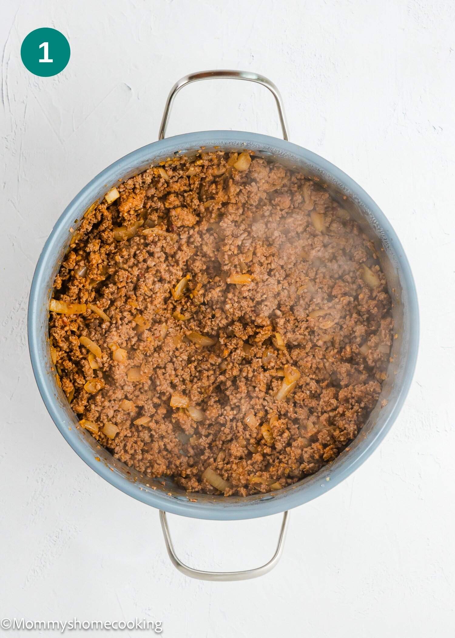 A large pan filled with cooked ground beef and onions, viewed from above on a white surface. Steam is rising from the mixture.