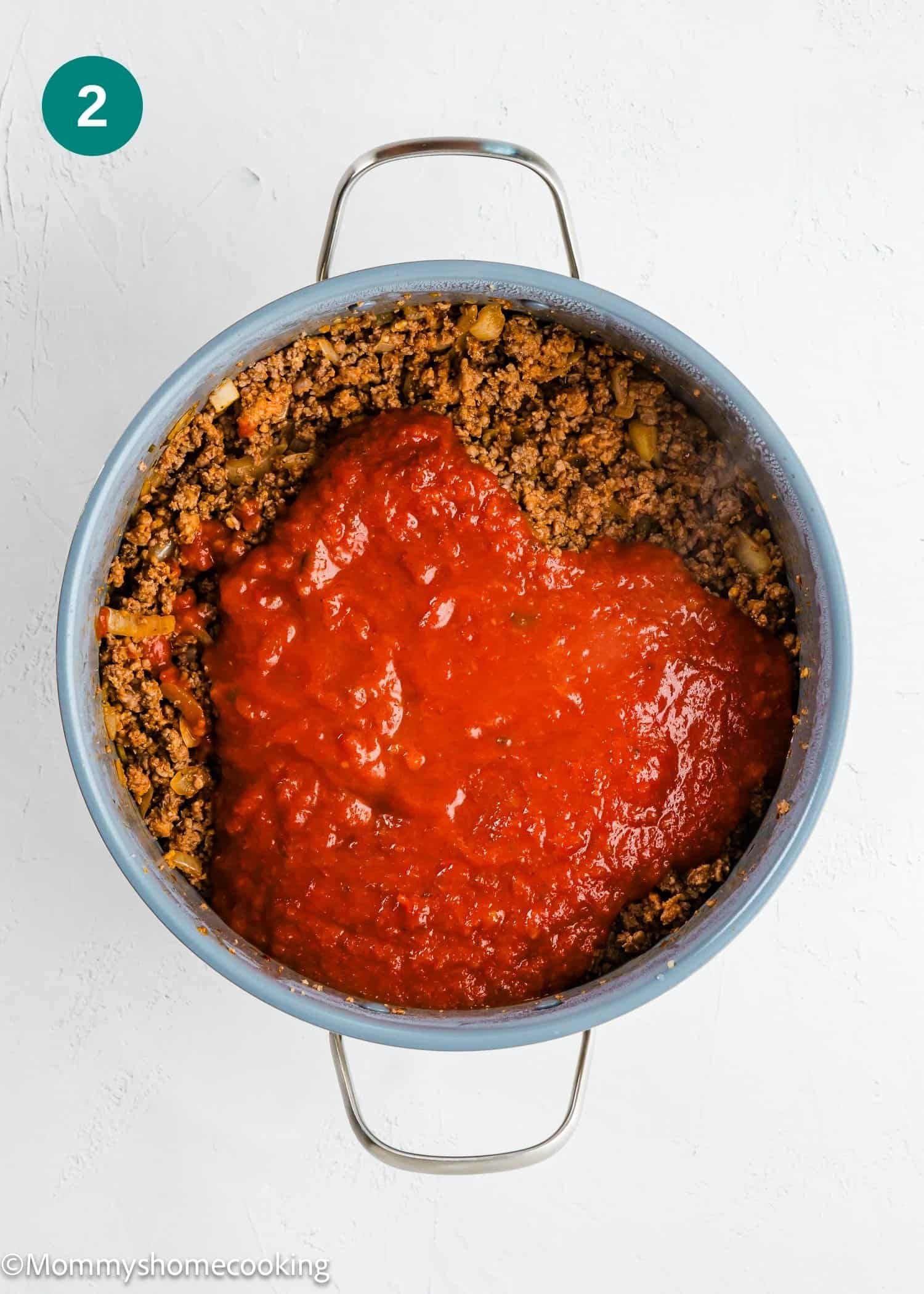 A pot containing cooked ground meat with tomato sauce poured on top, shown on a white surface.