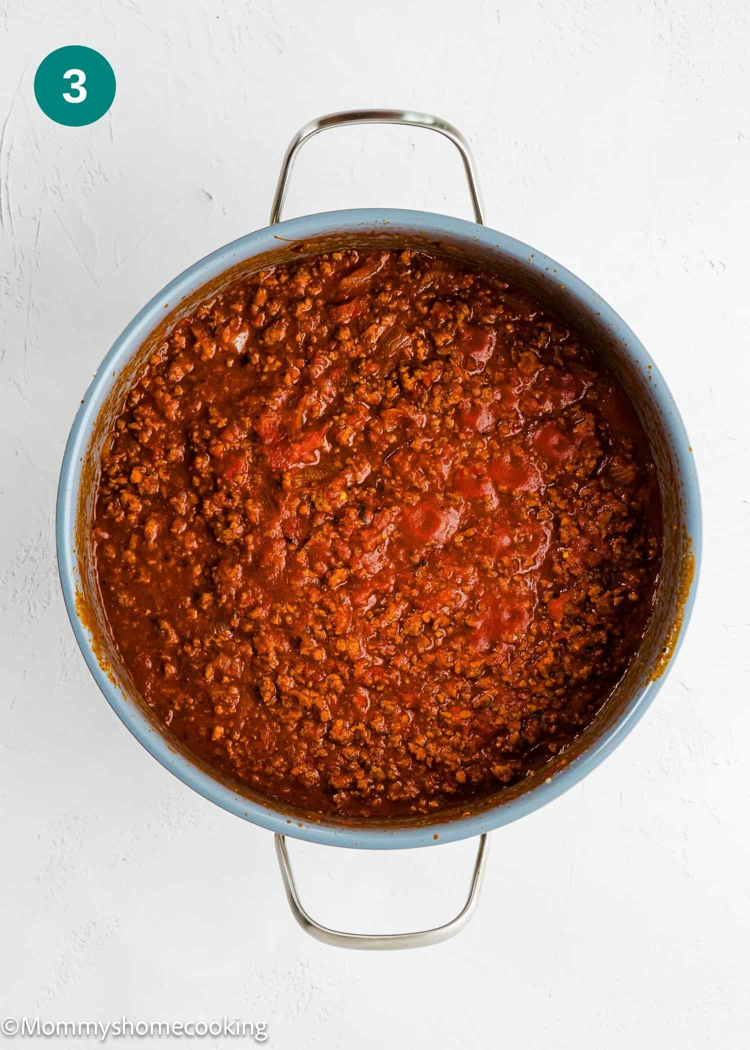 A pot filled with cooked ground meat in tomato sauce sits on a white surface, viewed from above.