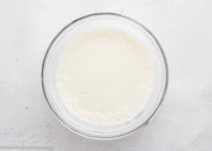 A glass bowl filled with a creamy white liquid mixture sits on a white textured surface.