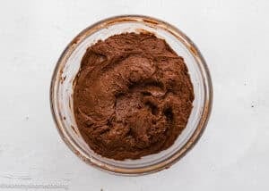 A clear glass bowl filled with thick chocolate batter sits on a white textured surface.