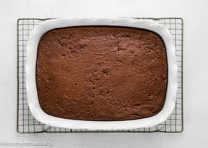 A rectangular chocolate cake baked in a white ceramic dish, cooling on a wire rack against a white background.