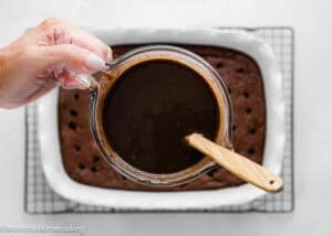 A hand holds a glass measuring cup filled with chocolate sauce above a baked cake with holes in it, ready for pouring.