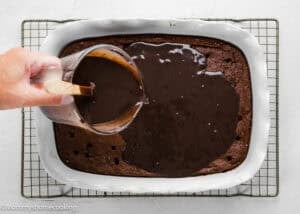 A hand pours chocolate sauce from a measuring cup over a baked chocolate cake in a white rectangular dish on a cooling rack.