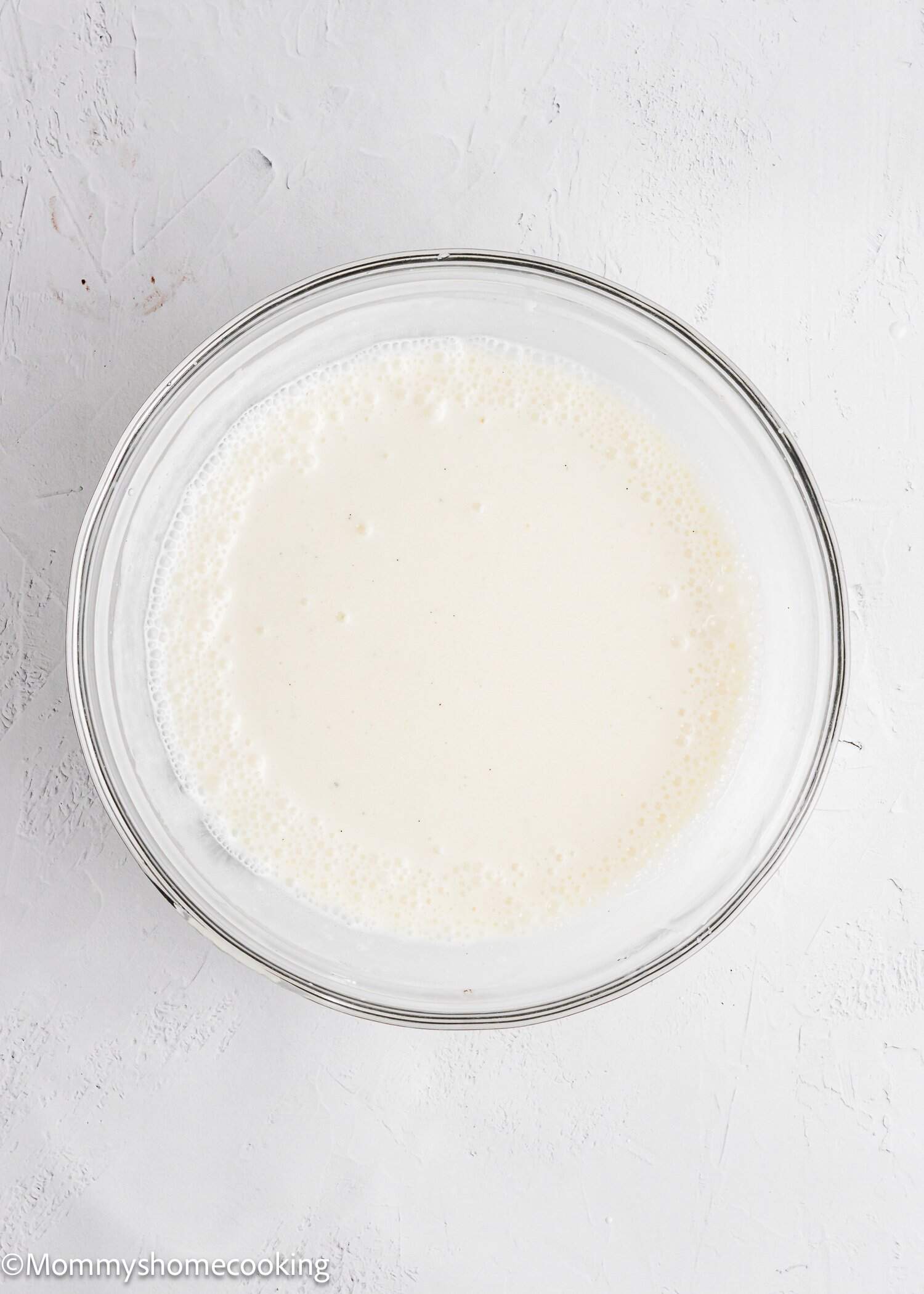 A glass bowl filled with a smooth, pale batter sits on a white textured surface.