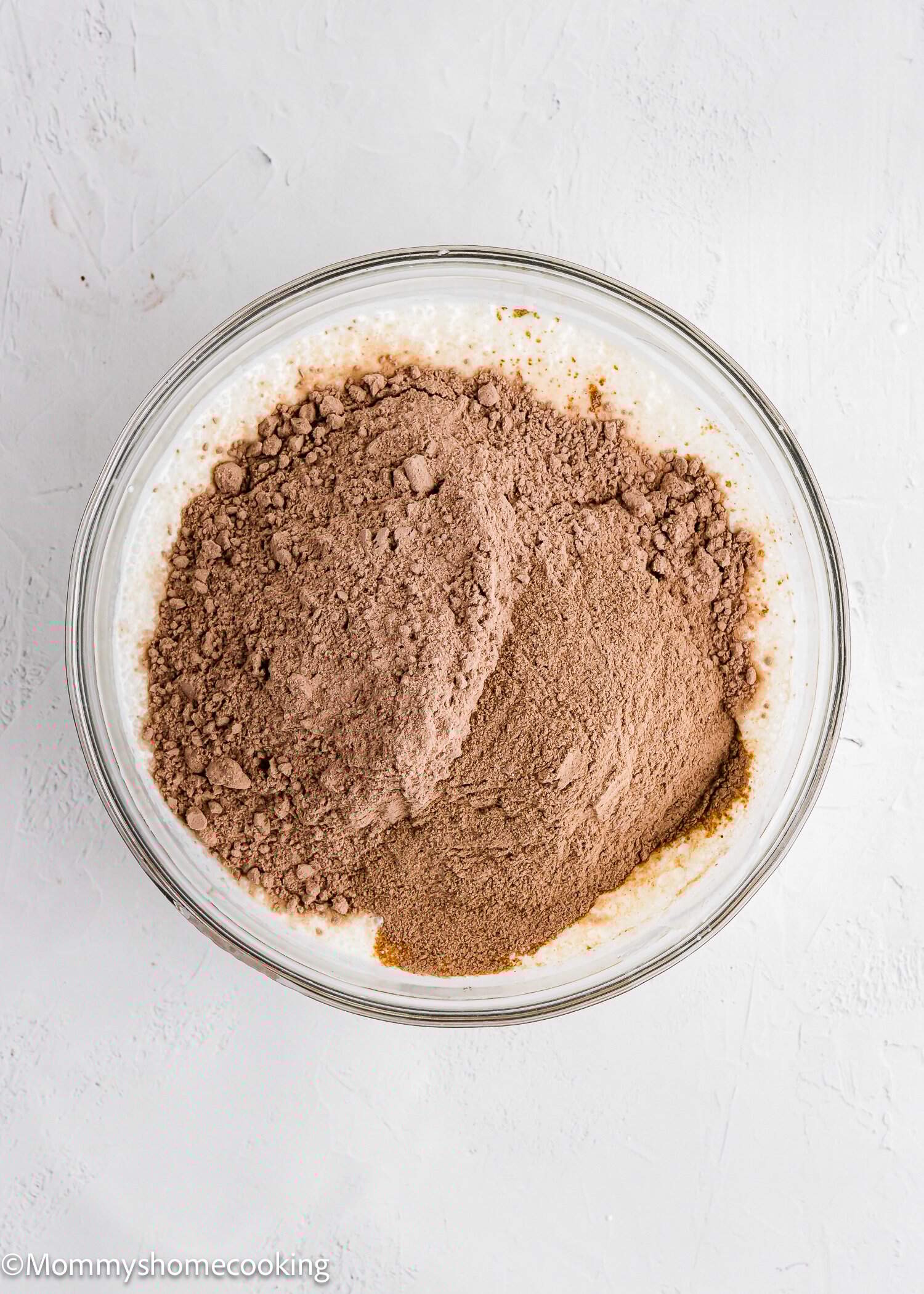 A glass bowl with a mixture of cocoa powder and white batter on a light surface.