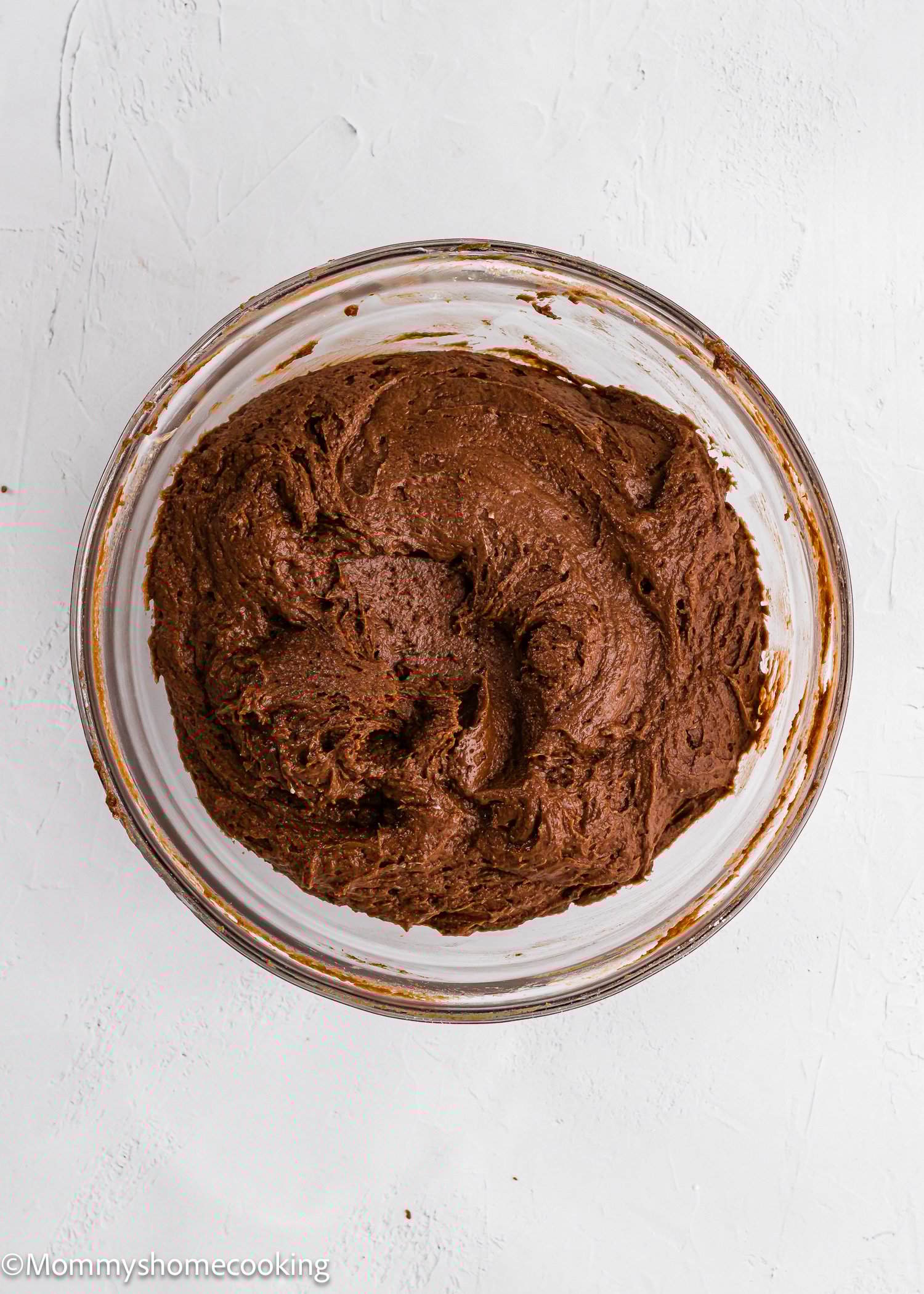 A glass bowl filled with thick, chocolate cake or brownie batter sits on a white surface.