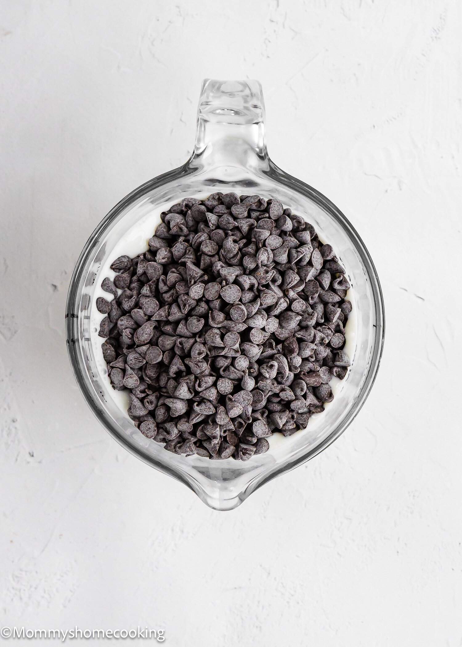 A glass measuring cup filled with chocolate chips sits on a white textured surface.