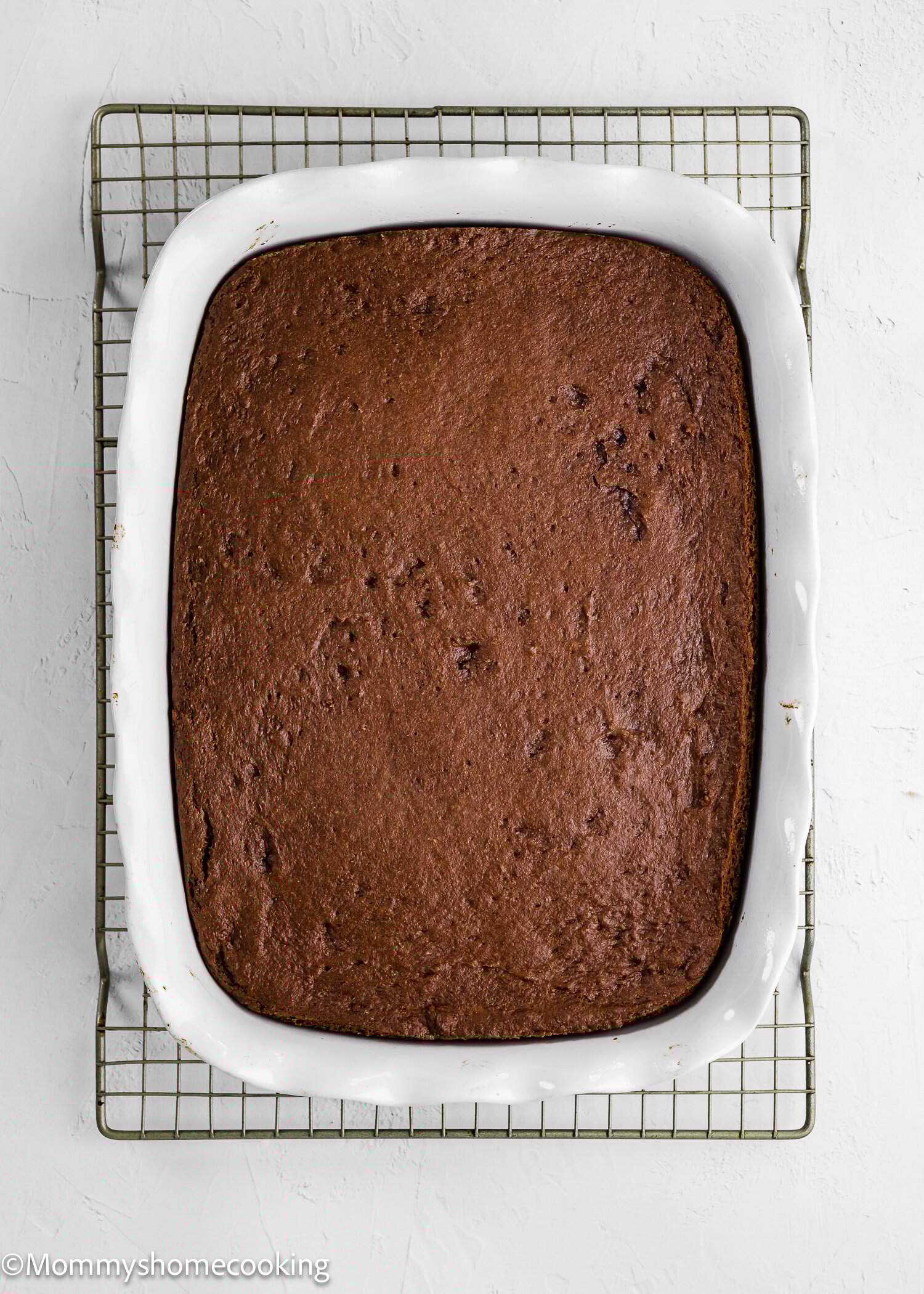 A rectangular chocolate cake in a white baking dish sits on a metal cooling rack against a white background.