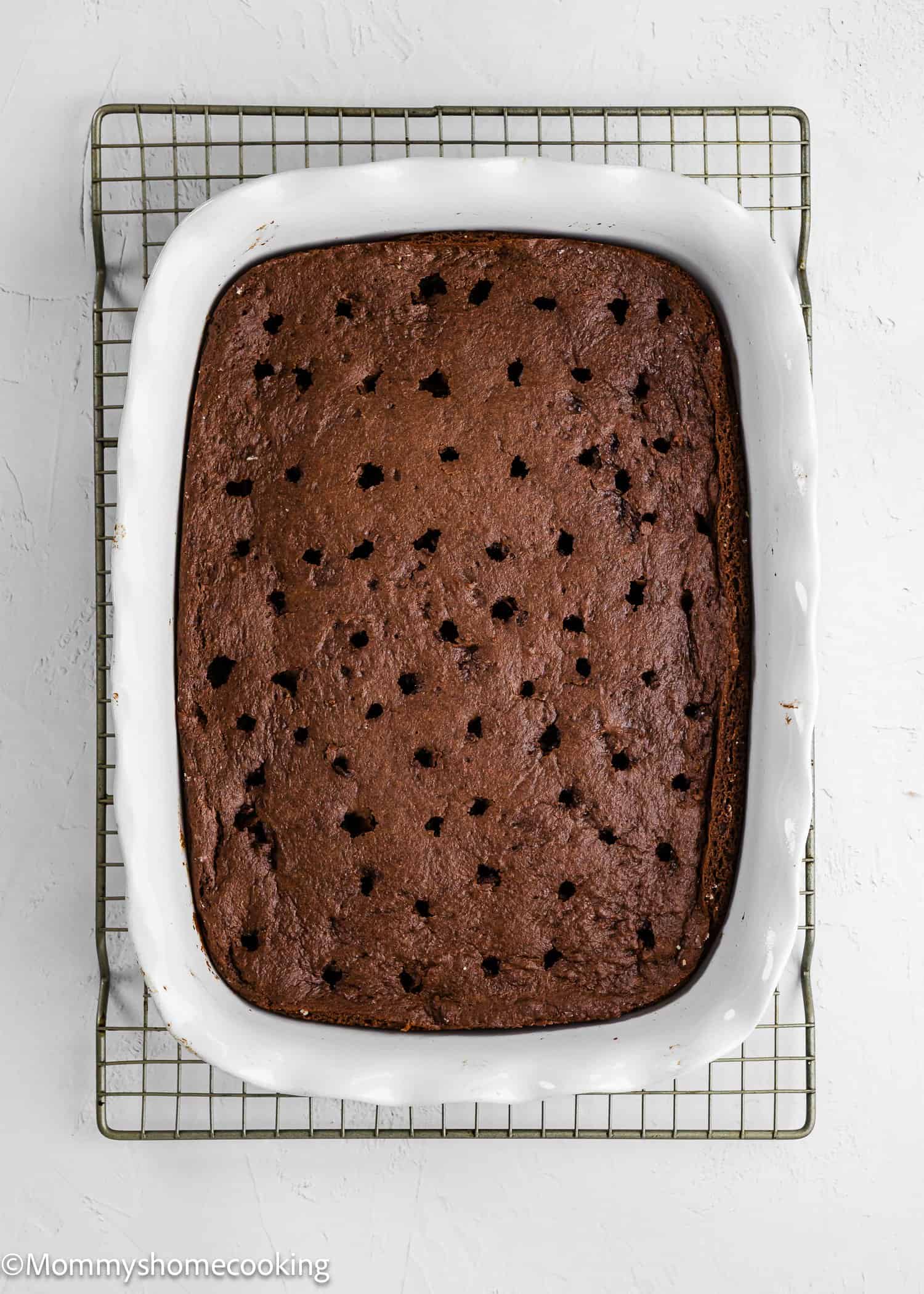 A rectangular chocolate cake with evenly spaced holes on top, baked in a white ceramic dish on a wire cooling rack.