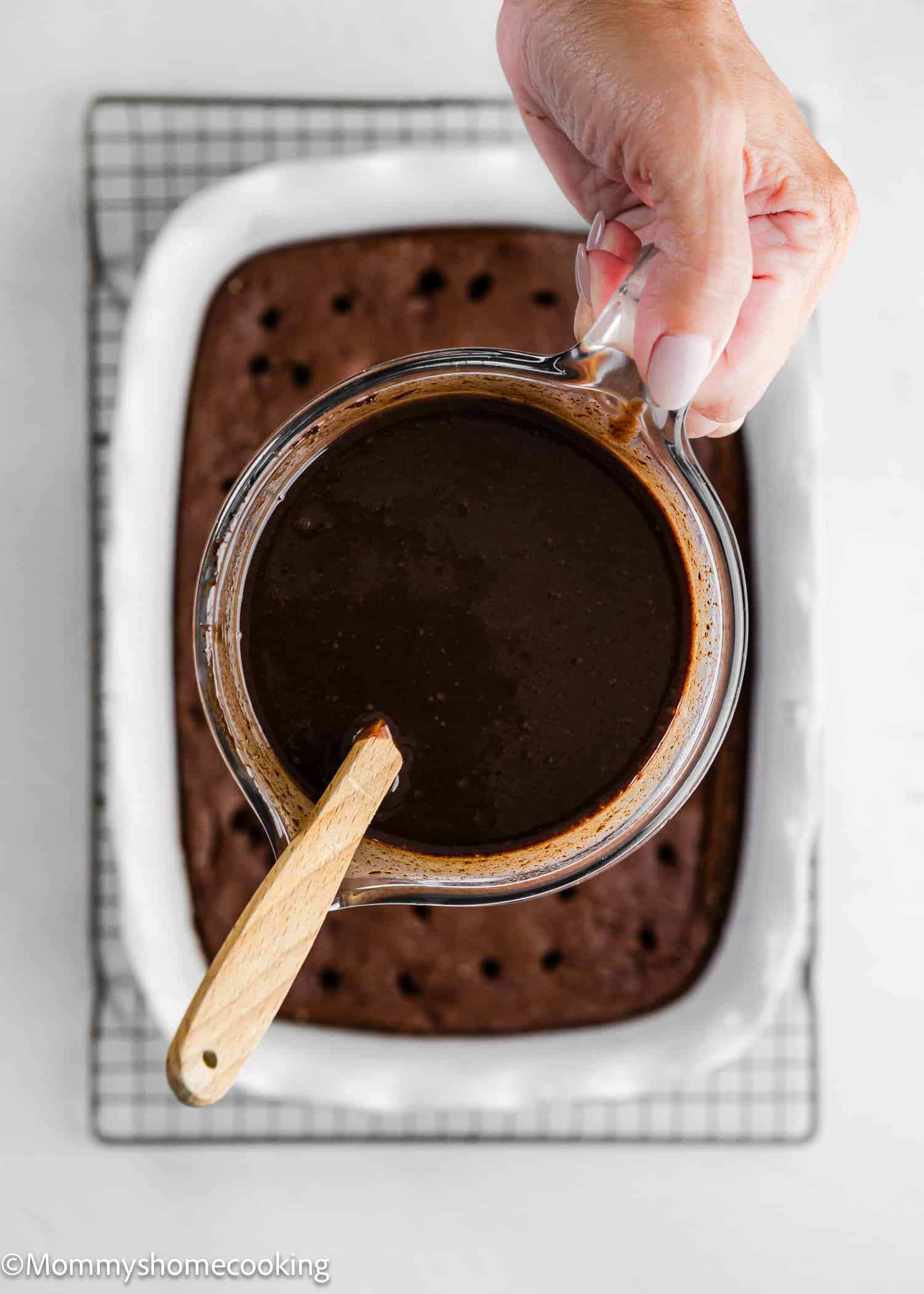 A hand holds a glass pitcher of chocolate sauce with a wooden spoon above a rectangular chocolate cake in a white baking dish.