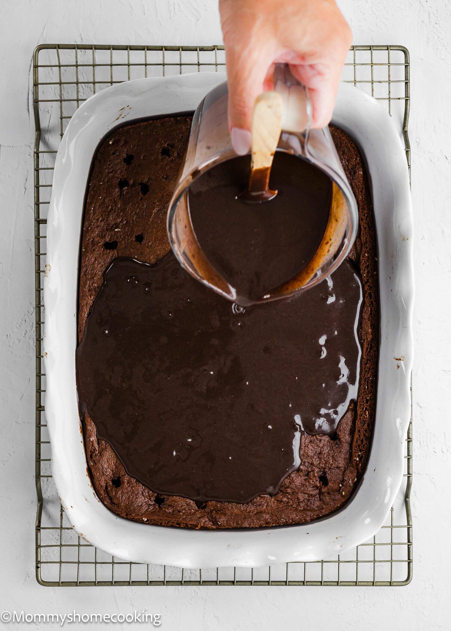 A hand pours chocolate sauce over a baked chocolate cake in a white rectangular dish placed on a cooling rack.