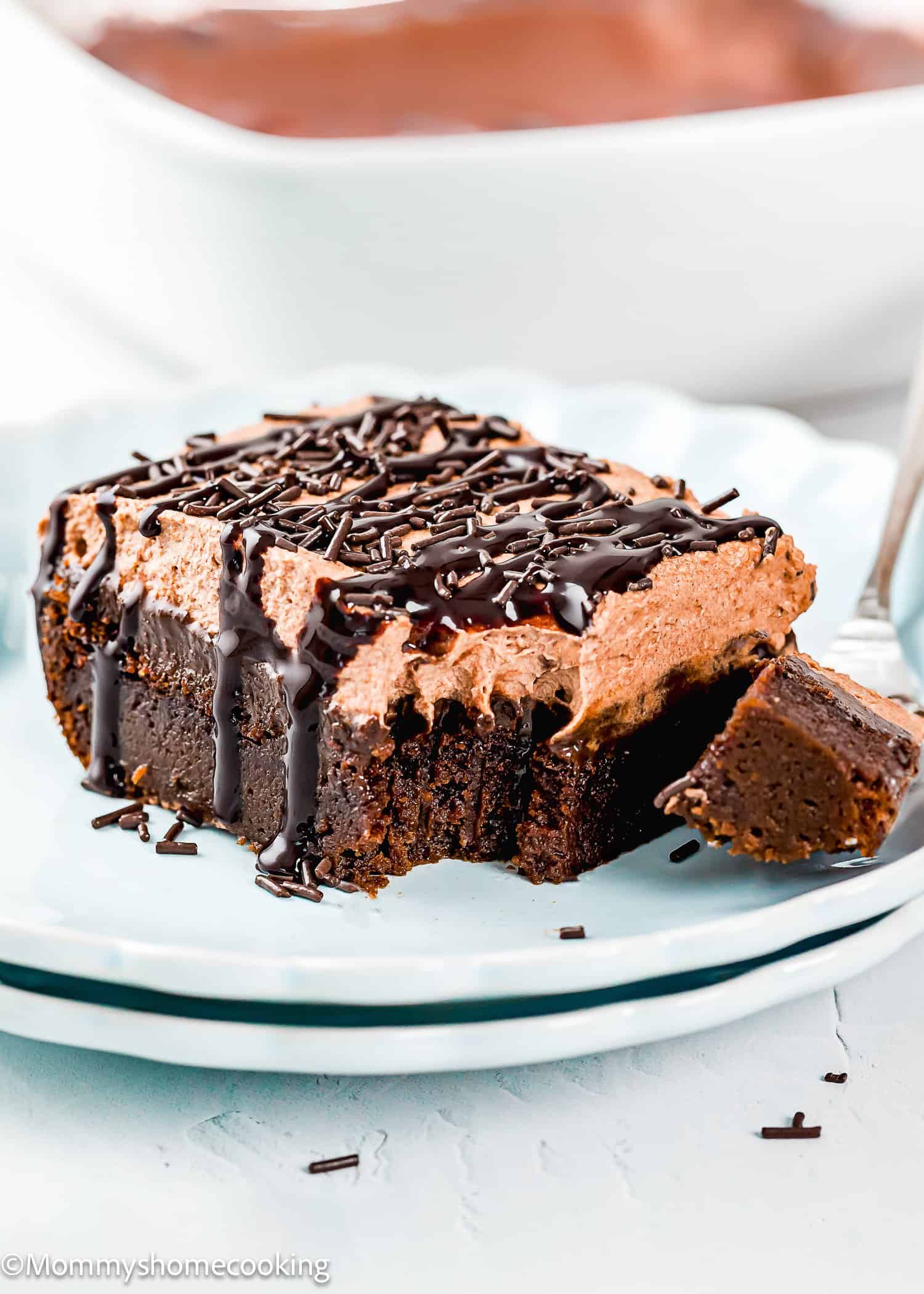 A slice of chocolate mousse cake topped with chocolate sprinkles and sauce sits on a plate, with a fork holding a small bite.