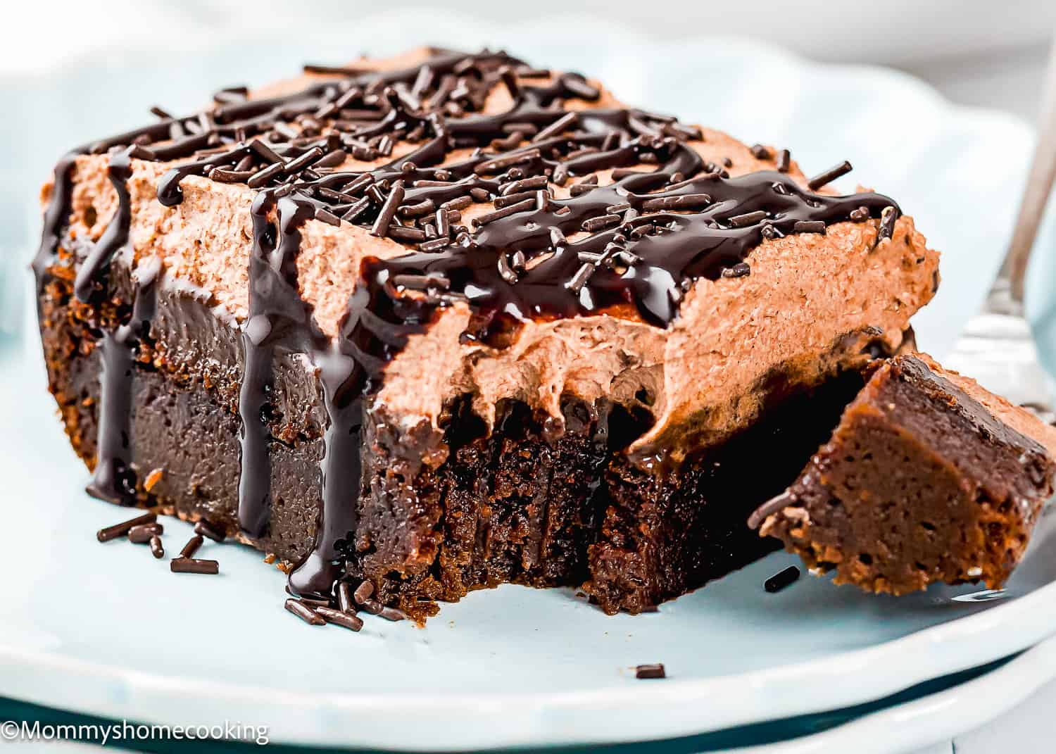 A slice of chocolate cake with chocolate frosting, chocolate sprinkles, and chocolate syrup on a white plate. A fork has taken a bite from the cake.