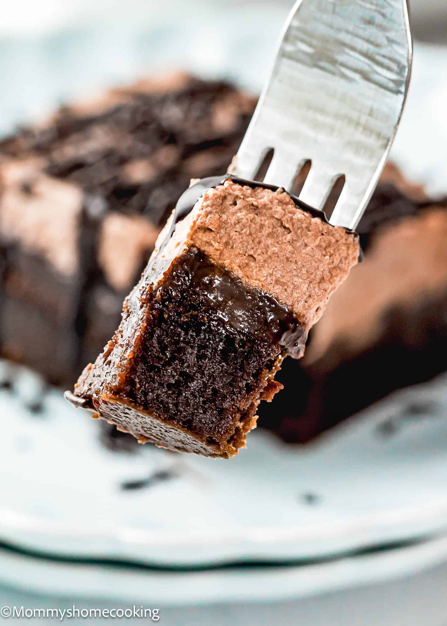 A close-up of a fork holding a bite of Fudgy Chocolate Pudding Poke Cake with a rich, gooey center and chocolate frosting, with the cake slice on a plate in the background.
