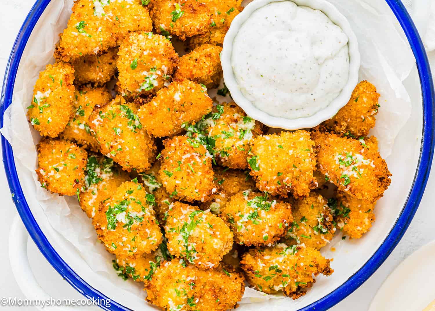 A bowl of golden-brown breaded and baked mushrooms, reminiscent of Garlic Parmesan Chicken Bites, garnished with chopped herbs and served with a small dish of creamy white dipping sauce.
