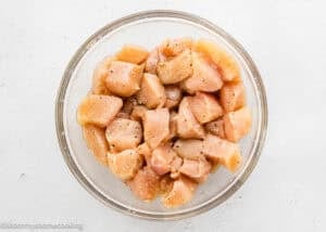A glass bowl filled with raw, diced chicken breast pieces seasoned with black pepper, ready to be transformed into flavorful Garlic Parmesan Chicken Bites, set on a white surface.