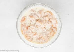 A glass bowl filled with raw Garlic Parmesan Chicken Bites marinating in a creamy, white herb sauce sits on a white textured surface.