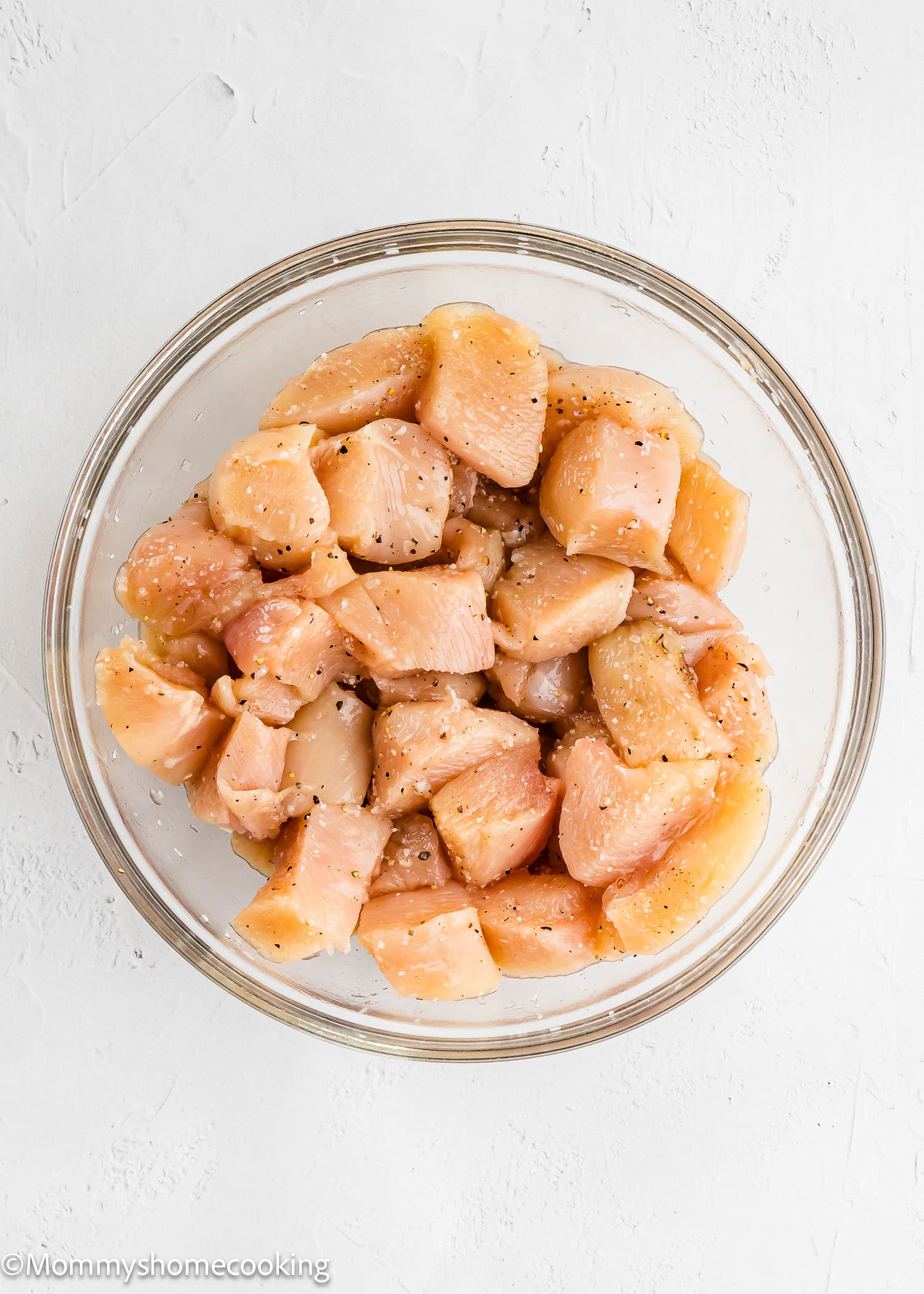 A glass bowl filled with raw, diced chicken breast pieces seasoned with salt and pepper, ready to be transformed into flavorful Garlic Parmesan Chicken Bites, sits on a white surface.