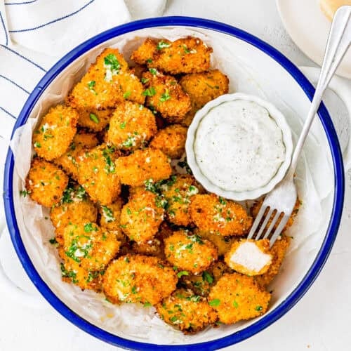 A bowl of crispy, breaded cauliflower bites garnished with herbs, served with a creamy dipping sauce. Reminiscent of Garlic Parmesan Chicken Bites, a fork holds one bite, and a striped towel is nearby.