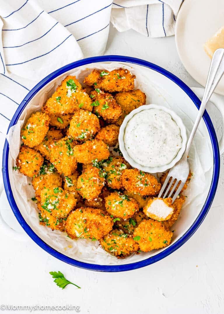 A bowl of crispy, breaded cauliflower bites garnished with herbs, served with a creamy dipping sauce. Reminiscent of Garlic Parmesan Chicken Bites, a fork holds one bite, and a striped towel is nearby.