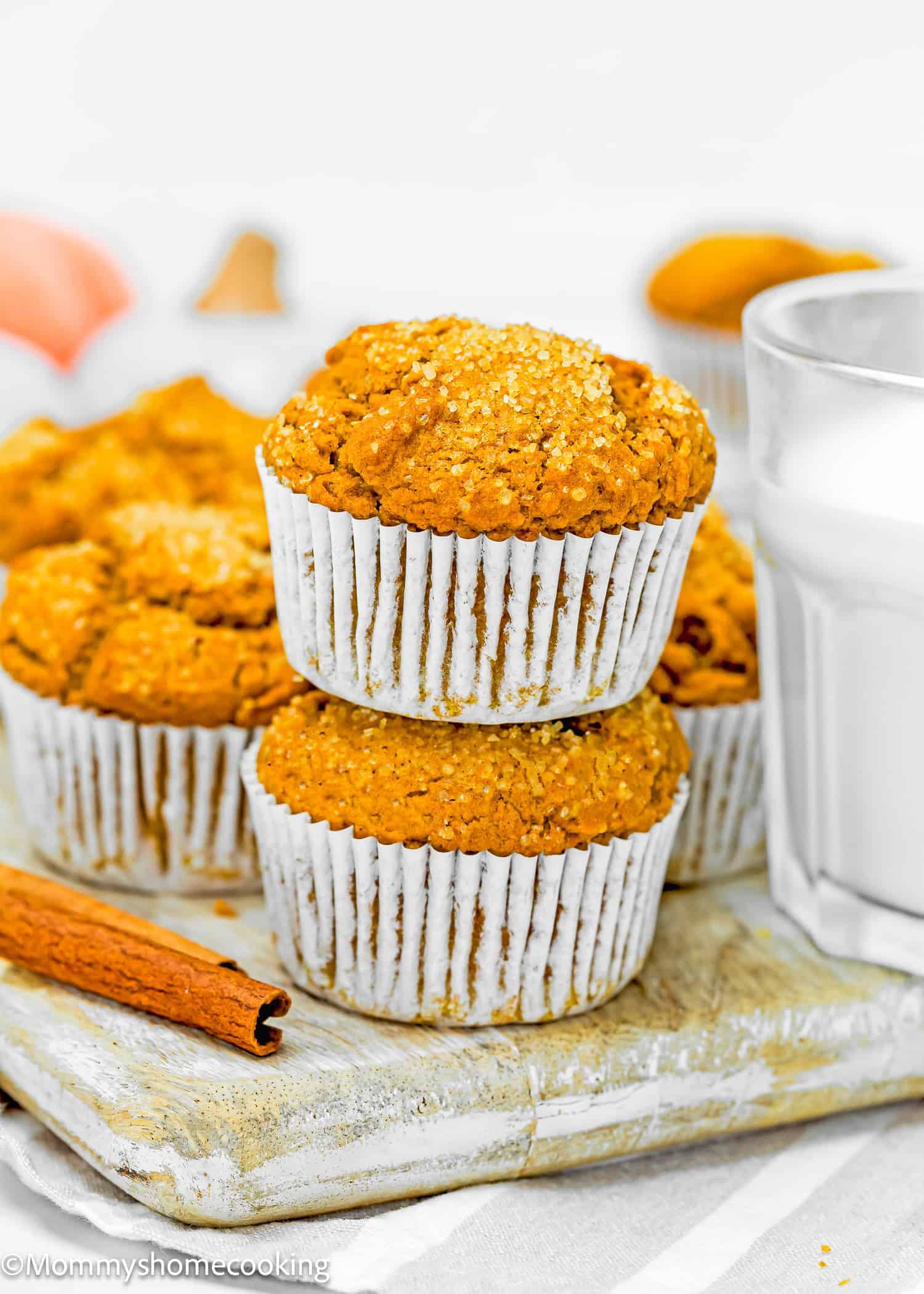 Two pumpkin muffins stacked on a wooden board next to a glass of milk and a cinnamon stick, with more muffins in the background.