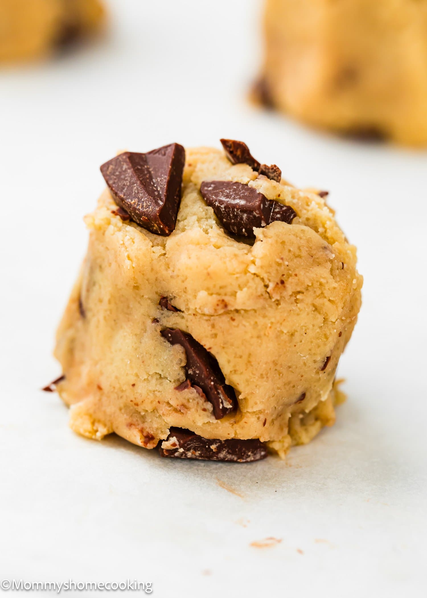 A close-up of a raw chocolate chip cookie dough ball sitting on a white surface, with visible chunks of chocolate embedded in the dough.