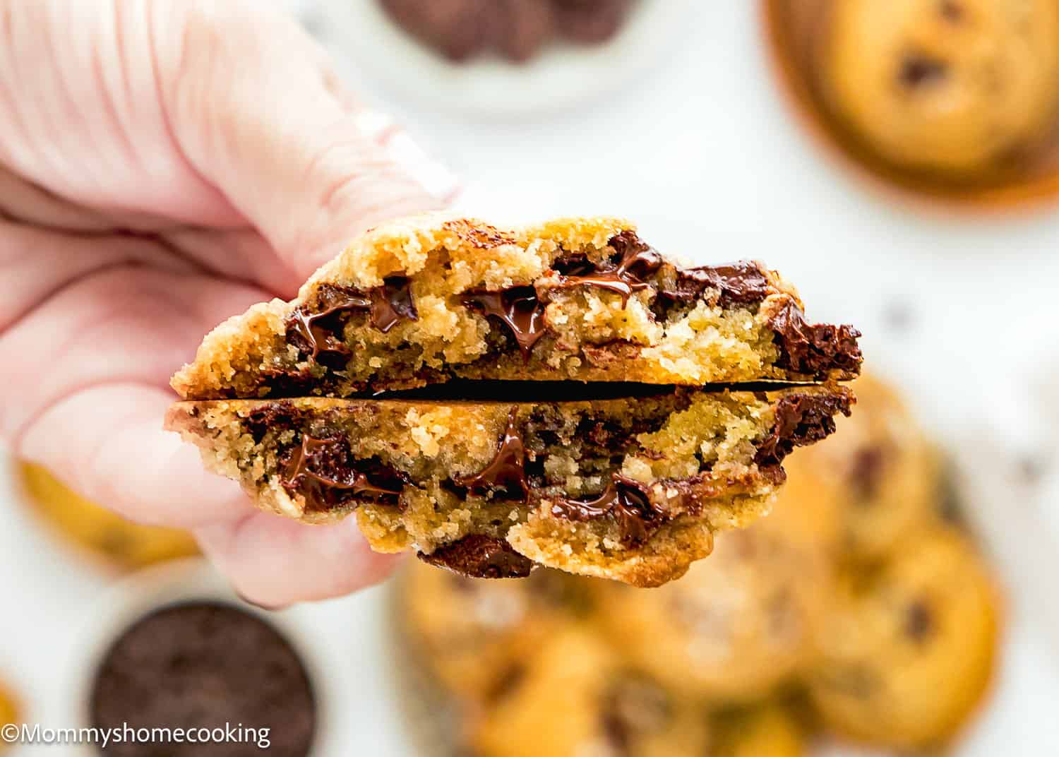 A hand holds a gooey chocolate chip cookie broken in half, revealing melted chocolate chips inside. Other cookies and chocolate chips are blurred in the background.