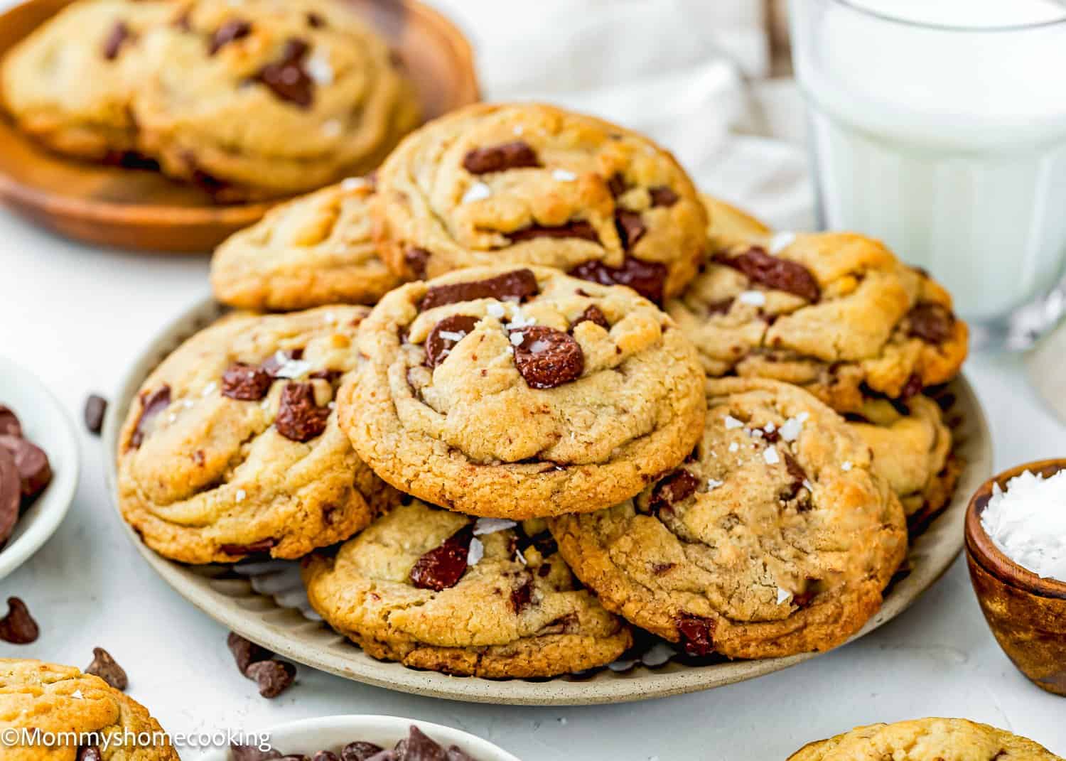 A plate of chocolate chip cookies topped with sea salt, surrounded by a glass of milk, a small bowl of chocolate chips, and a bowl of sea salt. The cookies look thick, chewy, and freshly baked.