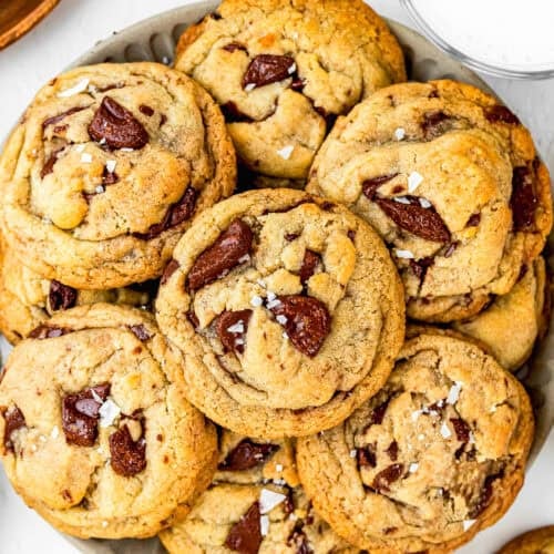 A plate of chunky egg-free chocolate chip cookies topped with sea salt, with a glass of milk and extra chocolate chips nearby. The cookies are golden brown and look soft and chewy.