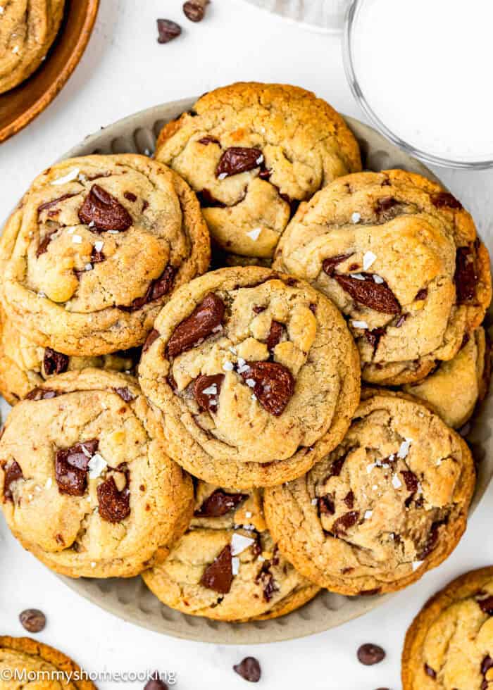 A plate of chunky egg-free chocolate chip cookies topped with sea salt, with a glass of milk and extra chocolate chips nearby. The cookies are golden brown and look soft and chewy.