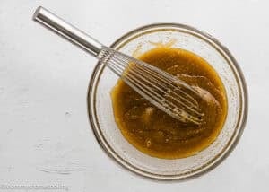 A metal whisk rests in a glass bowl filled with a thick, light brown mixture on a white surface.