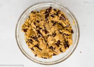 A clear glass bowl filled with chocolate chip cookie dough sits on a white surface. The dough is golden brown with visible chunks of dark chocolate mixed throughout.