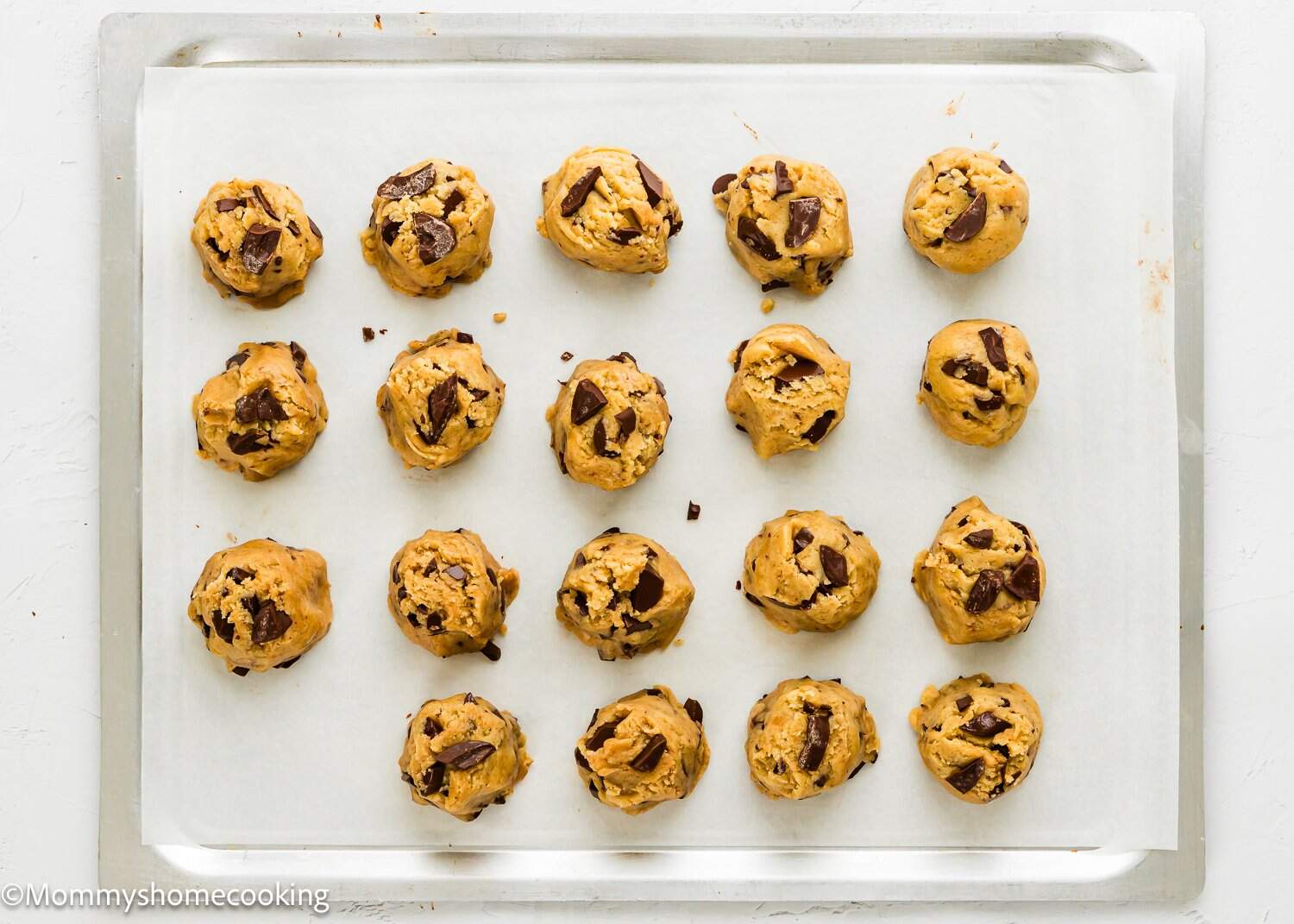 A baking tray lined with parchment paper holds 18 unbaked balls of chocolate chip cookie dough arranged in neat rows, ready to be baked.