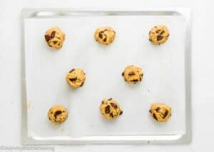 Nine unbaked chocolate chip cookie dough balls spaced evenly on a baking sheet lined with parchment paper, ready to be baked. The background is a clean, white surface.