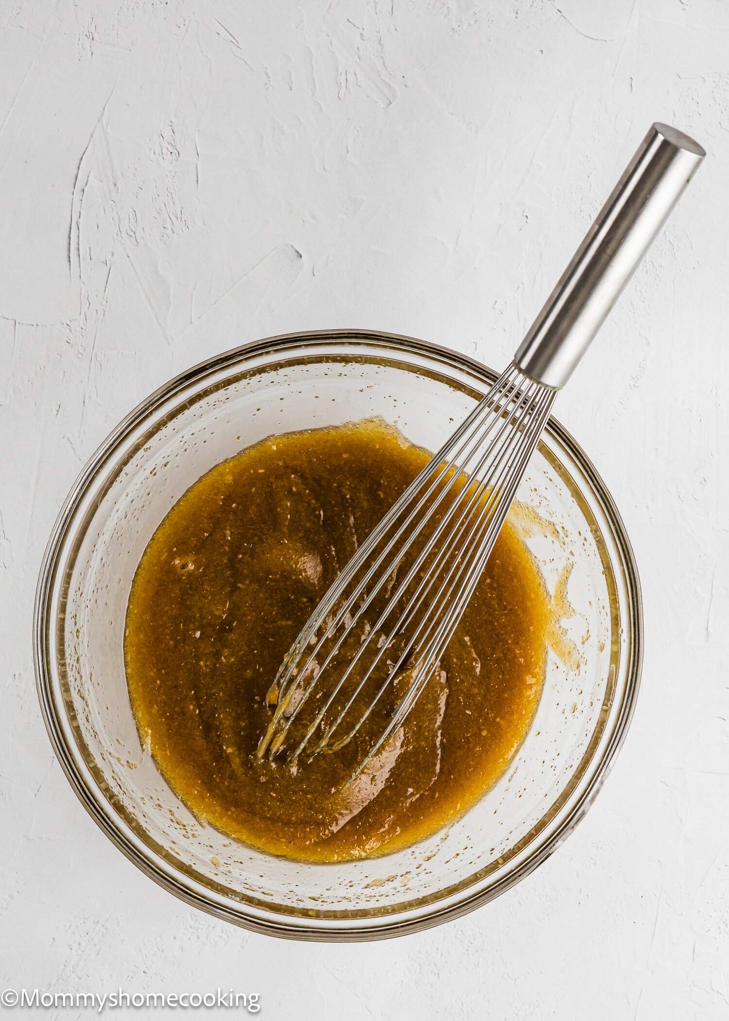 A glass bowl filled with a thick, brown liquid mixture being whisked with a metal whisk on a white textured surface.