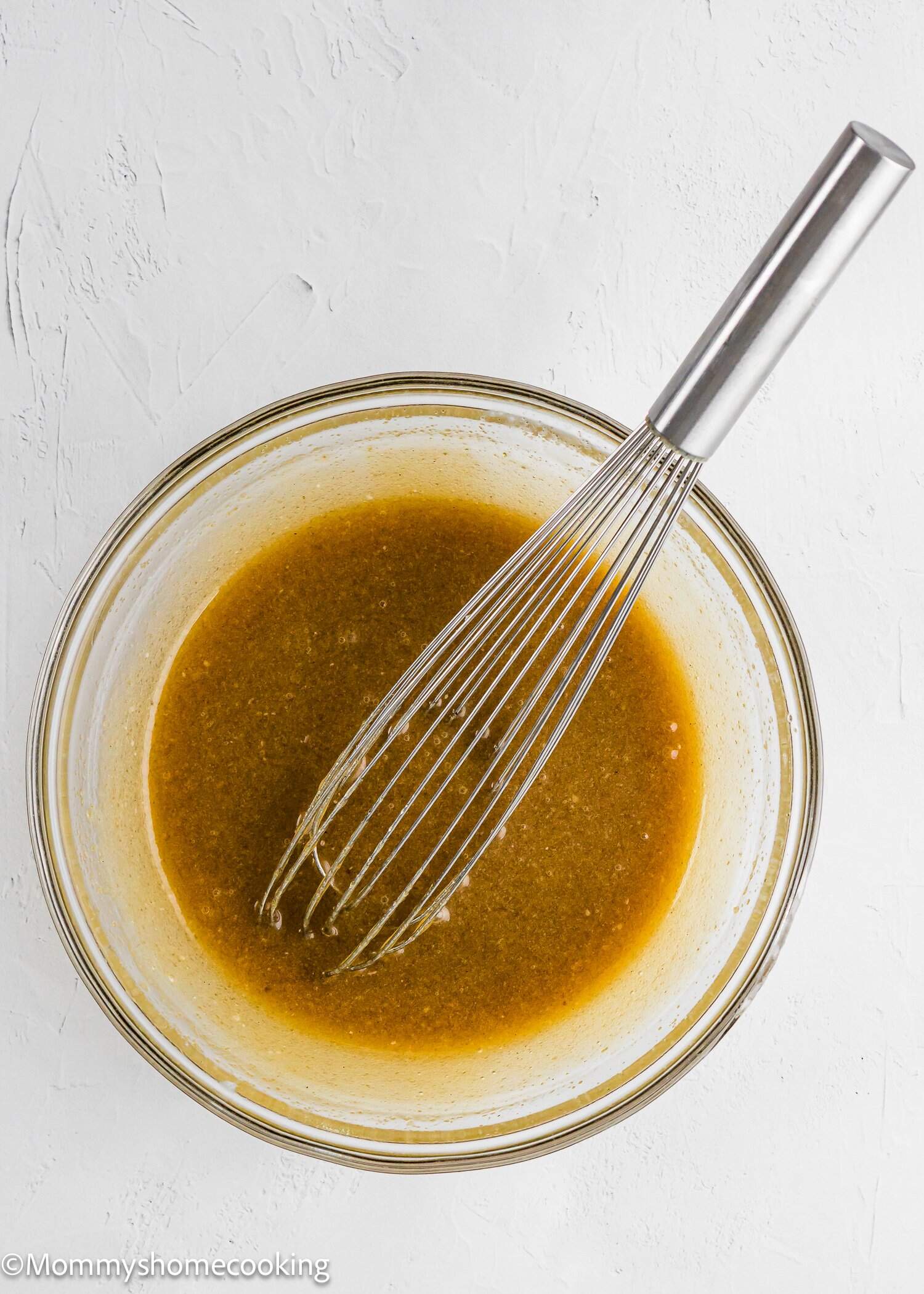 A glass bowl filled with a brownish liquid mixture and a metal whisk resting inside, placed on a white textured surface.