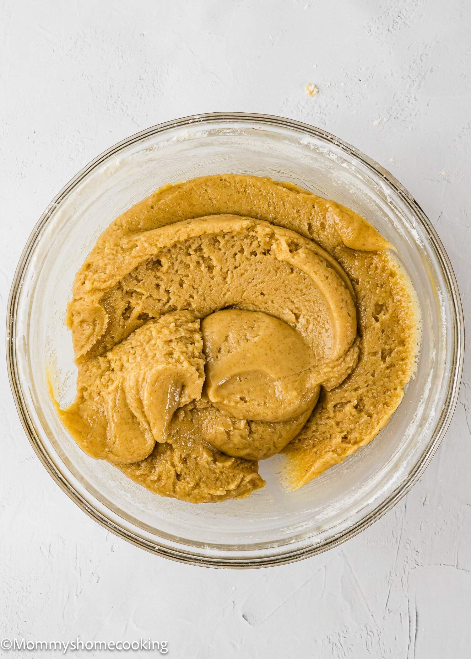 A glass bowl filled with smooth, light brown cookie dough sits on a white textured surface.