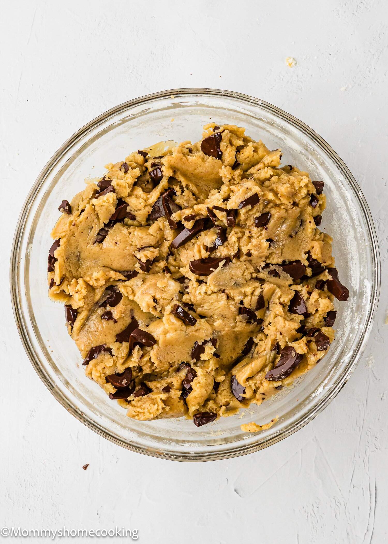 A clear glass bowl filled with chocolate chip cookie dough sits on a white surface. The dough is studded with large chunks of chocolate and appears soft and ready to be baked.