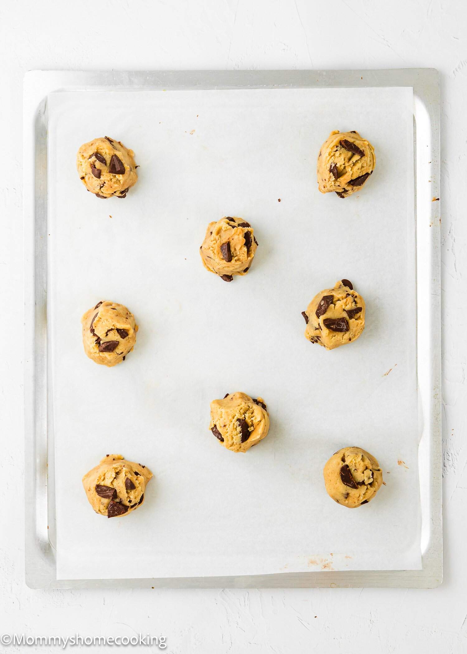 A baking sheet lined with parchment paper holds nine evenly spaced scoops of chocolate chip cookie dough, ready to be baked. The background is a white surface.