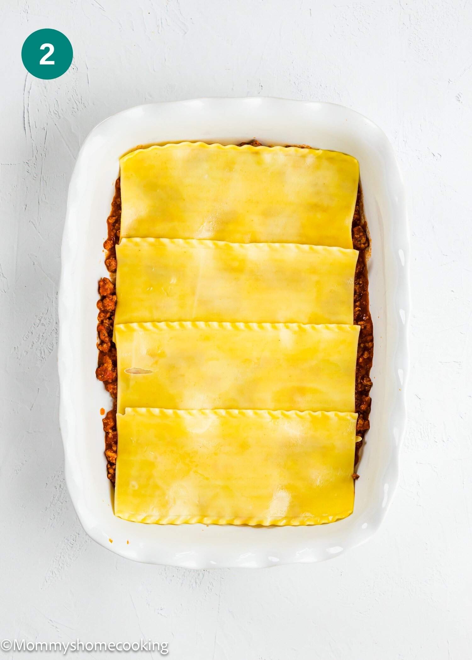 A white baking dish with a layer of meat sauce topped with four uncooked lasagna noodles.