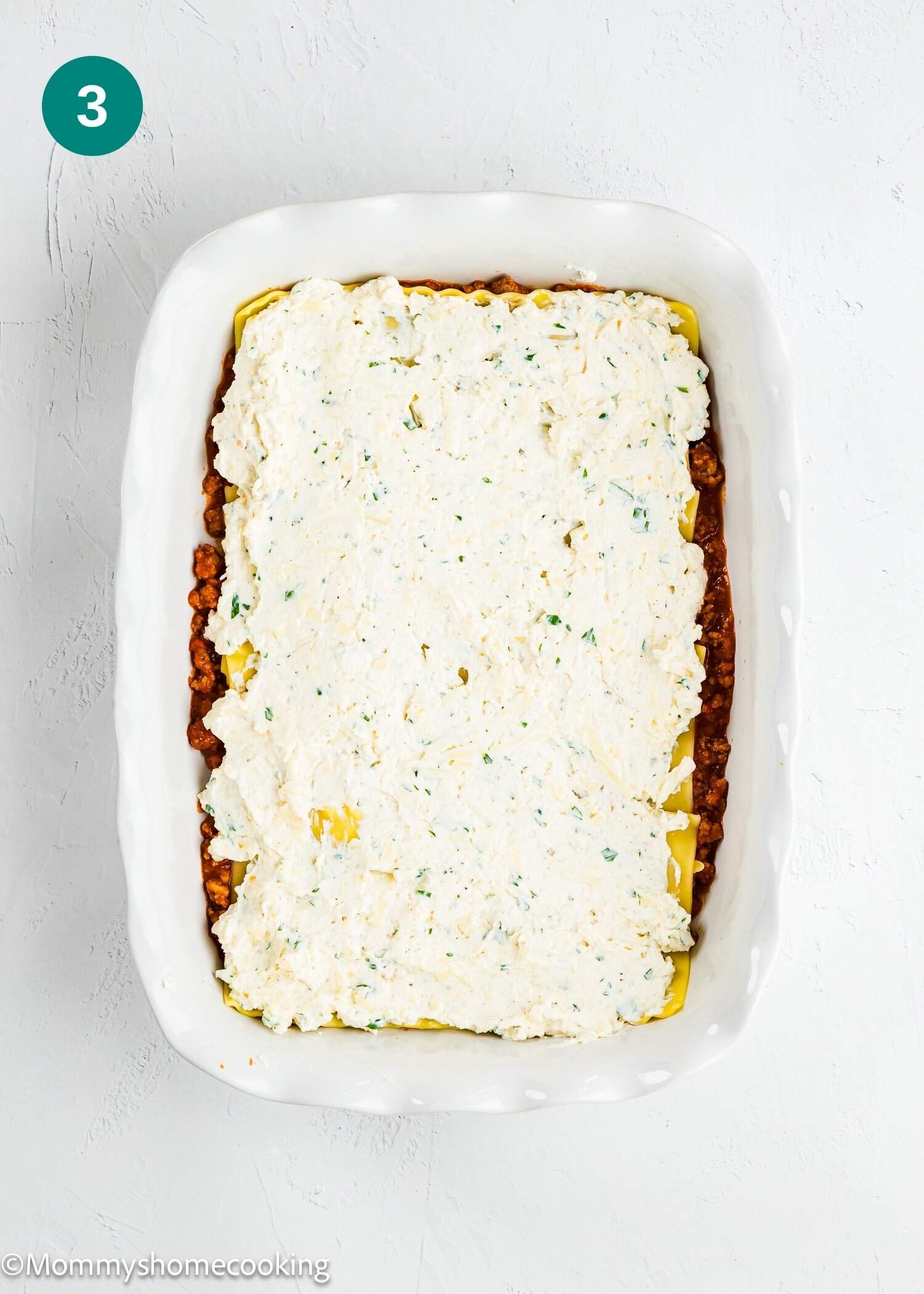 A rectangular baking dish with a layer of lasagna noodles topped with a spread of ricotta cheese mixture over a layer of meat sauce.