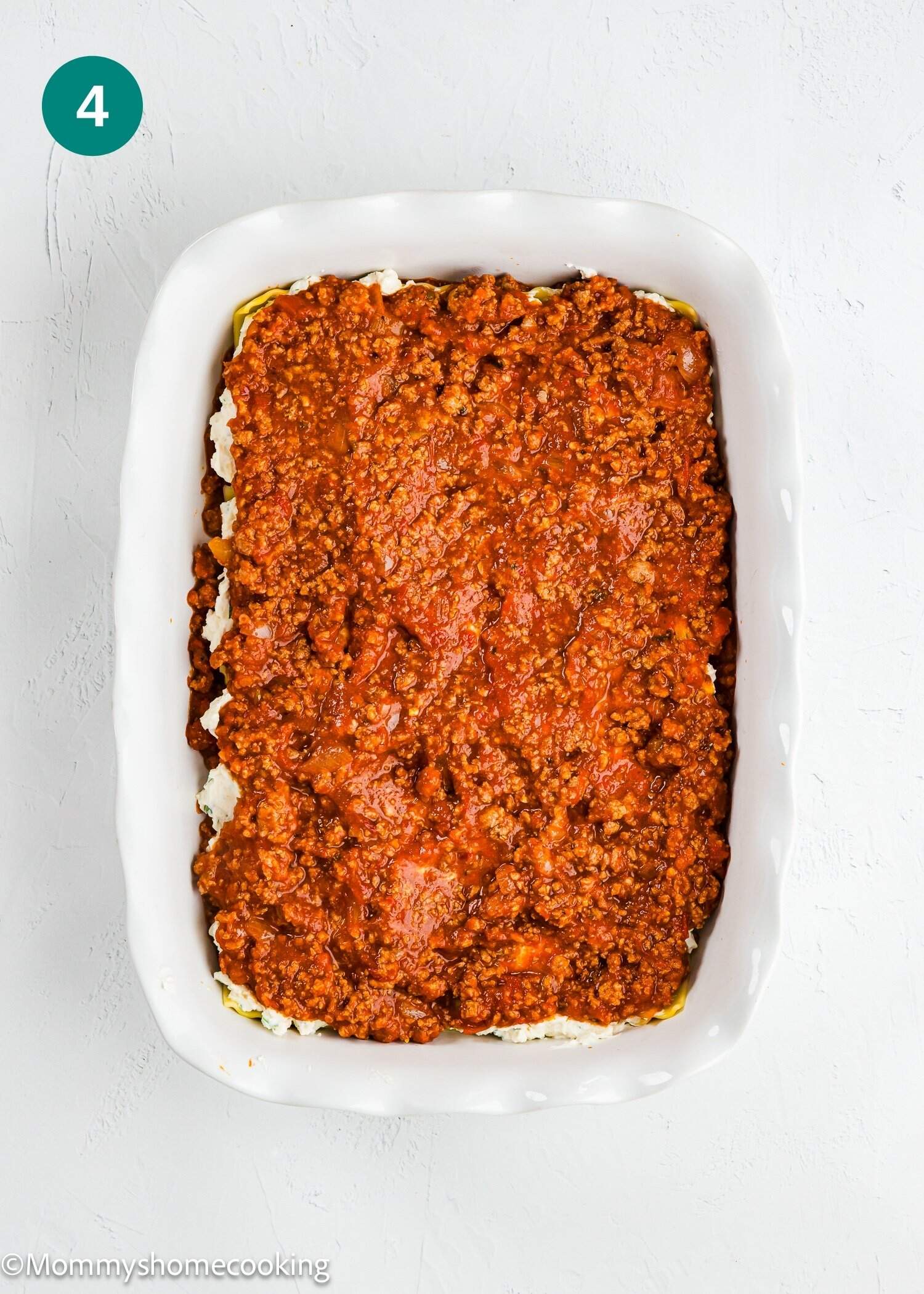 Rectangular white baking dish filled with a layer of ground meat tomato sauce mixture on top of a lighter layer, on a white surface. Step 4 indicator in the top left corner.