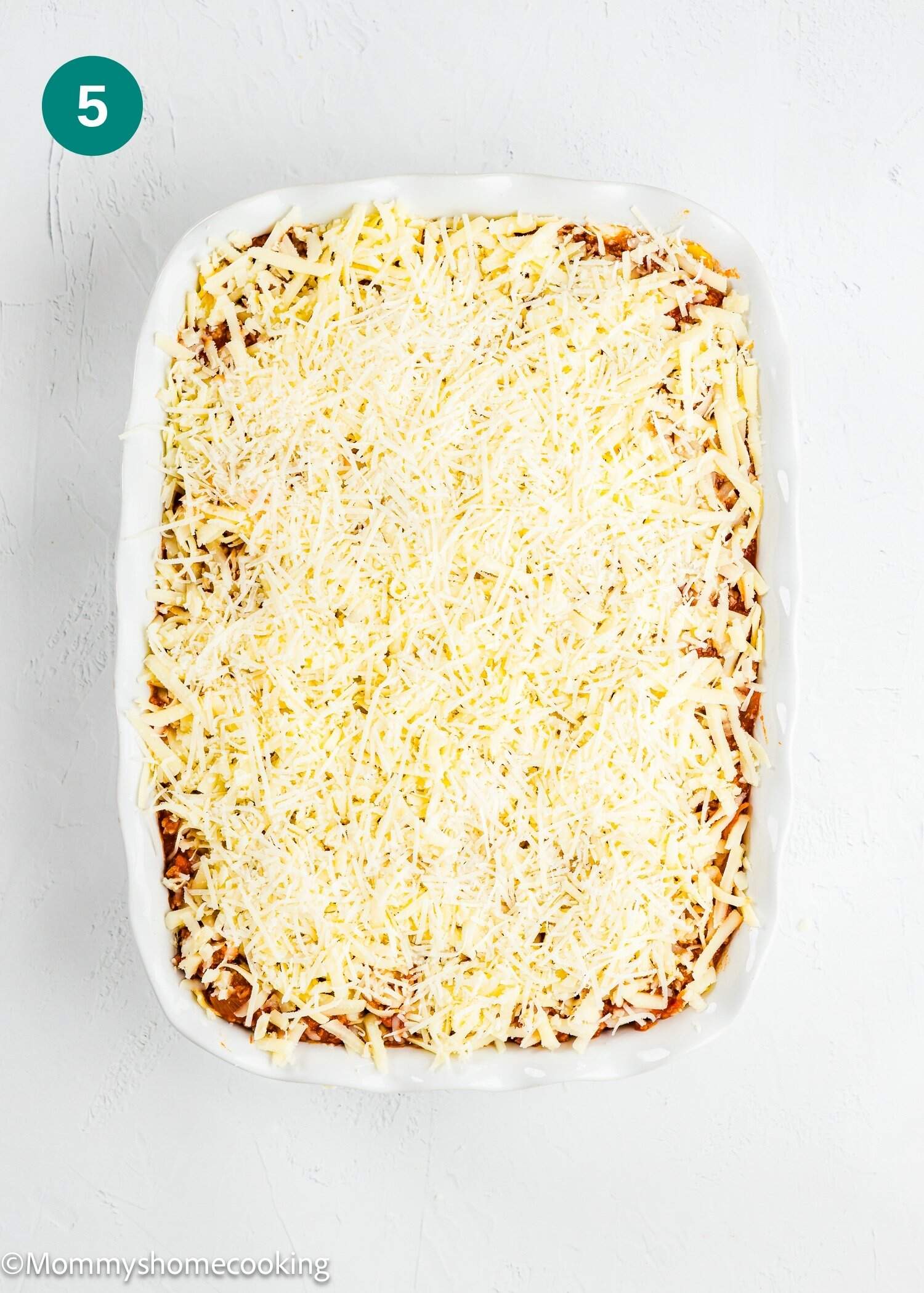 A white baking dish filled with a layered casserole topped with a generous amount of shredded cheese, ready to be baked.