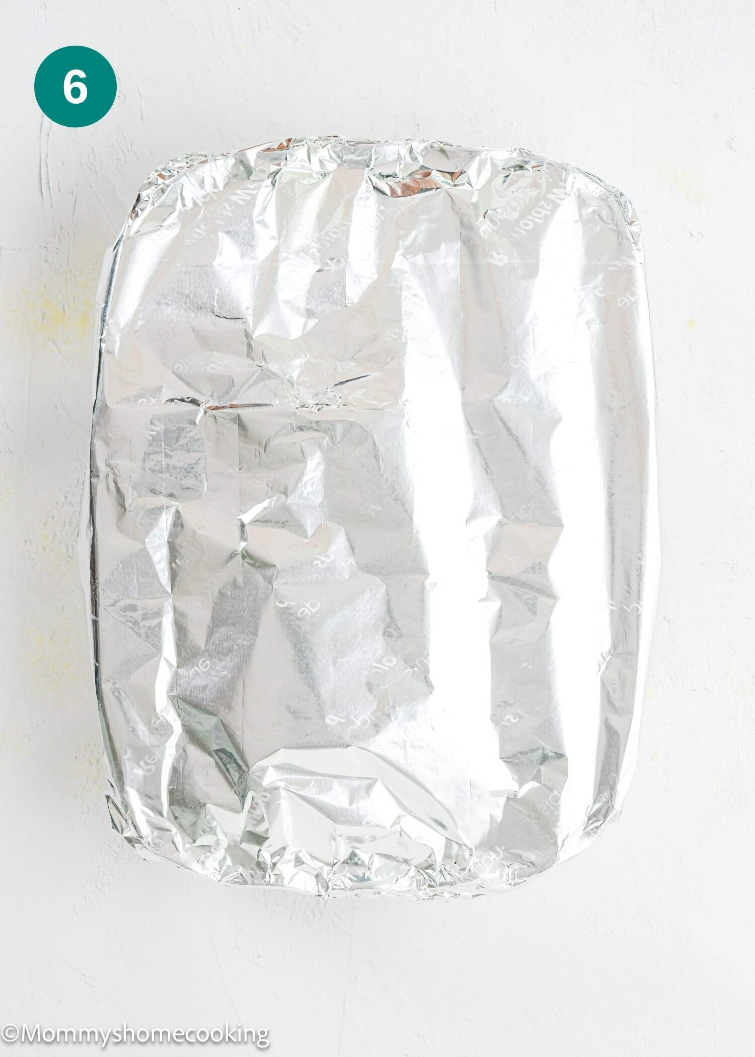 A rectangular baking dish covered tightly with aluminum foil, placed on a white surface. A green circle with the number 6 is in the upper left corner.