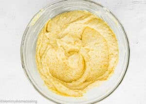 A glass bowl filled with yellow, creamy Easy Homemade Corn Pudding without Eggs batter sits on a white surface.